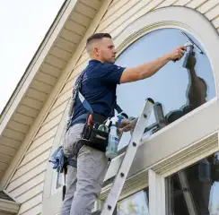 Professional window cleaner on a ladder using a squeegee to clean an exterior arched window, Nashville, TN.